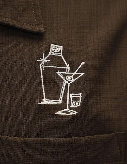 mørkebrune bowlingskjorte har et broderi af en drinkshaker, en martiniglas og et målebæger på venstre bryst, lige over brystlommen